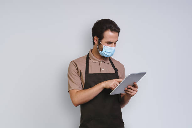 istock Handsome young man in apron wearing protective face and using digital tablet 1333947891