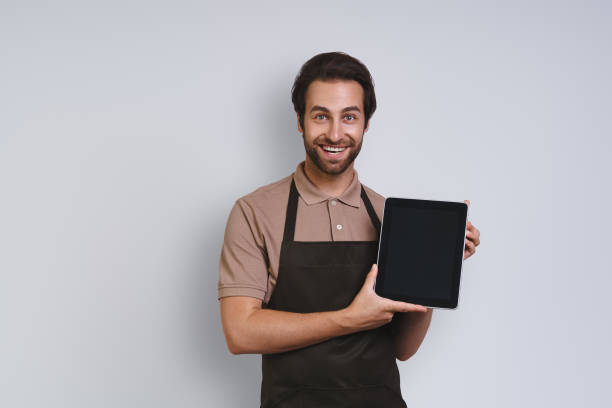 istock Cheerful young man in apron showing his digital tablet and smiling 1333948467