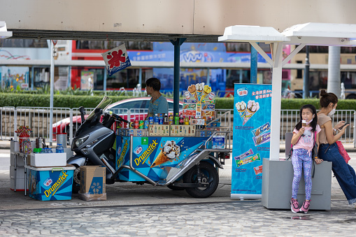 Hong Kong - August 13, 2021 : Ice cream and drinks vendor and a girl eating ice cream in Tsim Sha Tsui, Kowloon, Hong Kong.