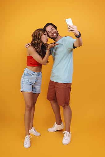 Full length of beautiful young smiling couple making selfie while standing against yellow background