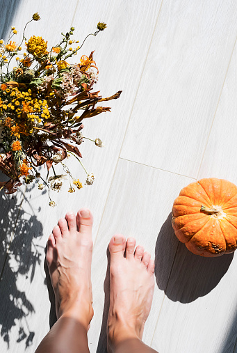 Autumn mood background. Barefoot woman legs, bouquet of dried flowers and pumpkin in sunlight.