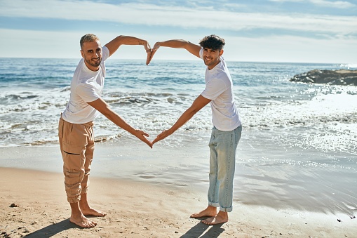 Young gay couple smiling happy doing heart symbol with arms at the beach.