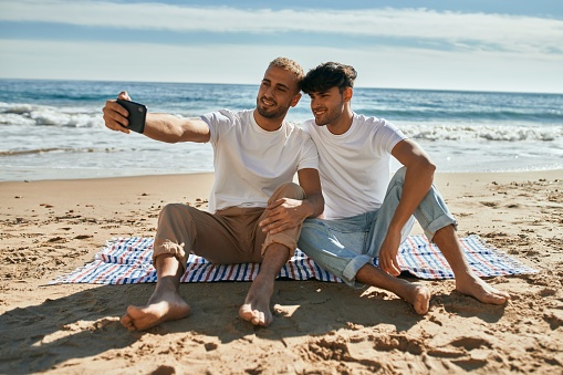 Young gay couple making selfie by the smartphone sitting at the beach.