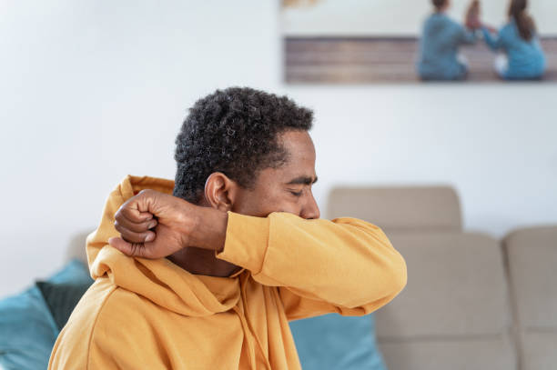 Adult man coughing or sneezing into elbow Adult man wearing a yellow hoodie in a living room, coughing or sneezing into elbow. coughing stock pictures, royalty-free photos & images