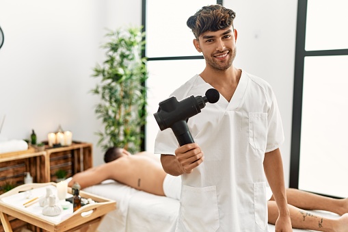 Young hispanic man holding therapy massage gun at wellness center looking positive and happy standing and smiling with a confident smile showing teeth