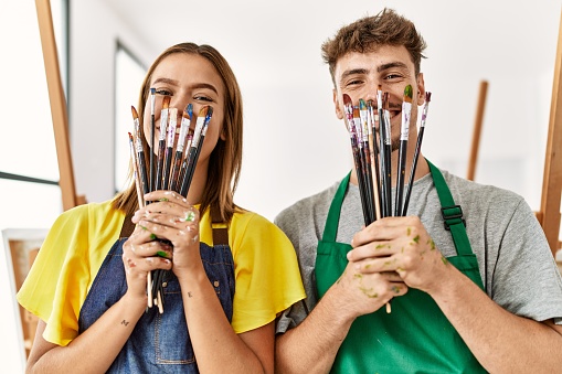 Young hispanic artist couple smiling happy covering mouth with paintbrushes at art studio.