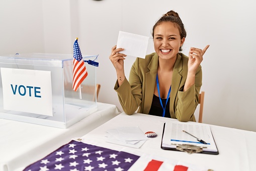 Beautiful hispanic woman holding voting envelop in ballot box smiling happy pointing with hand and finger to the side