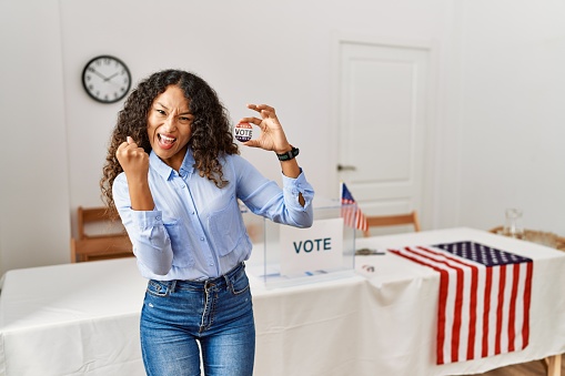 Beautiful hispanic woman standing by at political campaign by voting ballot very happy and excited doing winner gesture with arms raised, smiling and screaming for success. celebration concept.