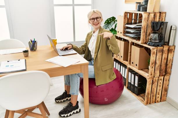 belle femme d’affaires caucasienne travaillant au bureau assise sur un ballon pilates pointant du doigt vers une auto-souriante heureuse et fière - yoga ball photos et images de collection