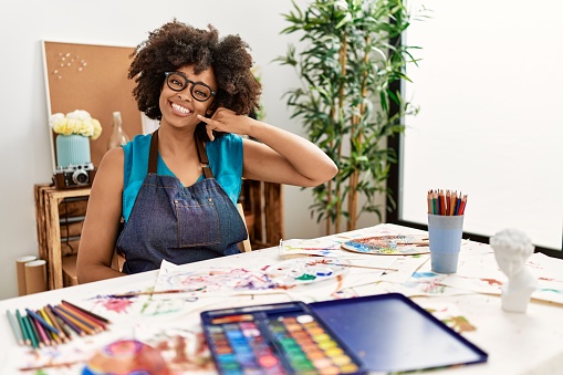 Beautiful african american woman with afro hair painting at art studio smiling doing phone gesture with hand and fingers like talking on the telephone. communicating concepts.