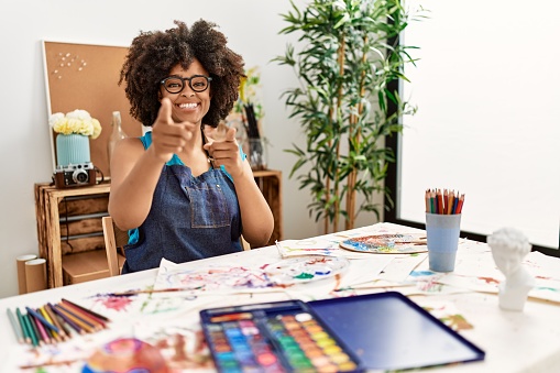 Beautiful african american woman with afro hair painting at art studio pointing fingers to camera with happy and funny face. good energy and vibes.