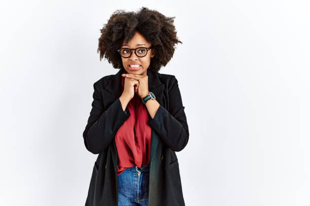 ビジネスジャケットと眼鏡をかけたアフロヘアのアフリカ系アメリカ人女性は、緊張して笑い、顎に手を向けて興奮した - blush ストックフォトと画像