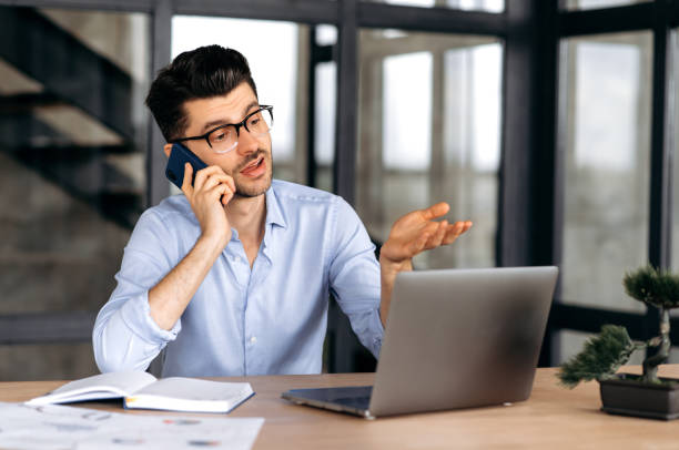 istock Confident Caucasian freelancer talking on smartphone while sitting at desk in modern office at Ð° laptop, successful man in strict shirt, have phone conversation, gesturing with his hand 1333969024