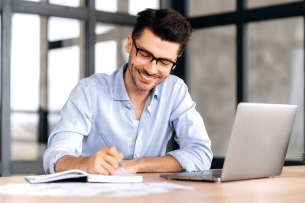 istock Smart successful caucasian man with glasses, sitting at the workplace, using a laptop, taking notes, studying or working online, listening to a webinar, gaining knowledge, smiling 1333969050