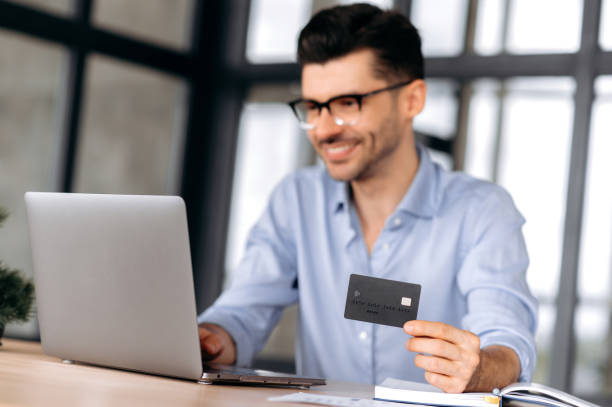 istock Blurred young modern Caucasian man wearing glasses, in casual clothes, sitting at table, using laptop and credit card to pay for online purchases or deliver goods home, smiling. Online shopping 1333969059