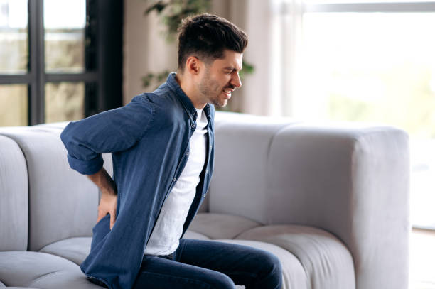 Sad upset young adult caucasian bearded man, in casual clothes, sitting on the sofa in the living room, massaging his back, suffering from back pain, suffering from arthritis, sciatica stock photo