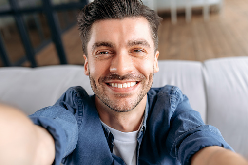 Young adult caucasian man takes a selfie. Portrait of attractive cheerful guy, in denim shirt, sitting on sofa, holding smartphone in hands and looking at webcam, smiling friendly