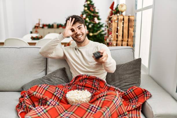 istock Young hispanic man eating popcorn sitting on sofa by christmas tree smiling happy doing ok sign with hand on eye looking through fingers 1333970648