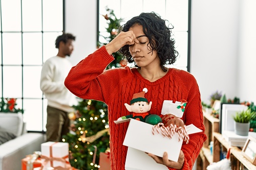 Young hispanic woman standing by christmas tree with decoration tired rubbing nose and eyes feeling fatigue and headache. stress and frustration concept.