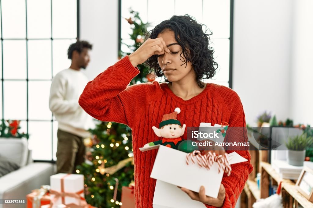 Young hispanic woman standing by christmas tree with decoration tired rubbing nose and eyes feeling fatigue and headache. stress and frustration concept. Emotional Stress Stock Photo