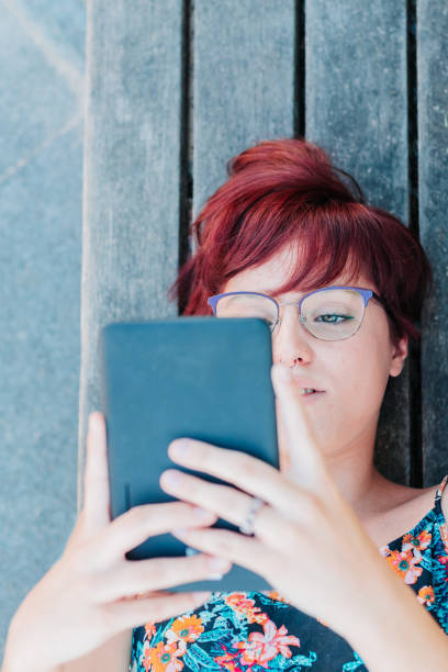 image verticale d’une jeune étudiante avec des lunettes lisant un livre électronique ou une tablette détendue à l’extérieur - women grass glasses e reader photos et images de collection