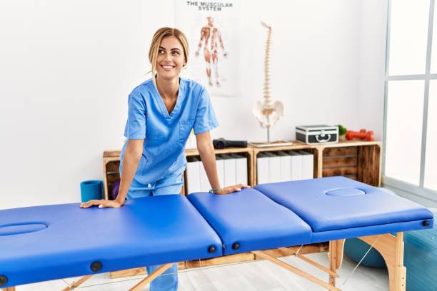 jovem fisioterapeuta caucasiano sorrindo feliz inclinando-se sobre a mesa na clínica - massage therapist - fotografias e filmes do acervo