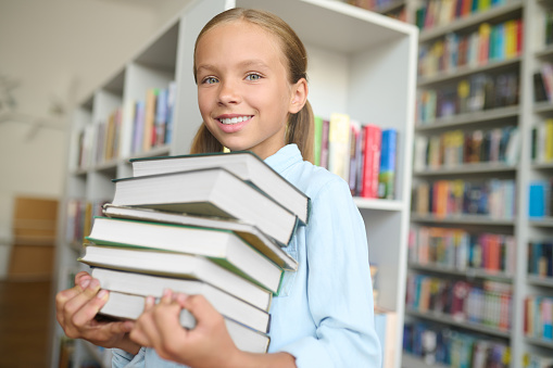 Waist-up portrait of a joyful cute pupil with a pile of books in her hands smiling at the camera
