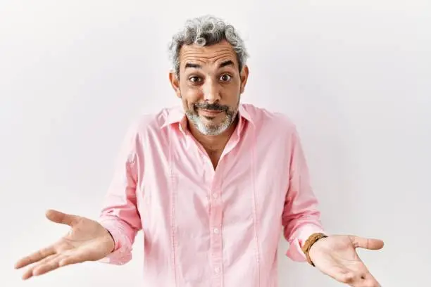 Photo of Middle age hispanic man standing over isolated background clueless and confused expression with arms and hands raised. doubt concept.