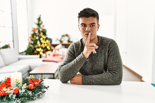Young hispanic man sitting on the table by christmas tree asking to be quiet with finger on lips. silence and secret concept.