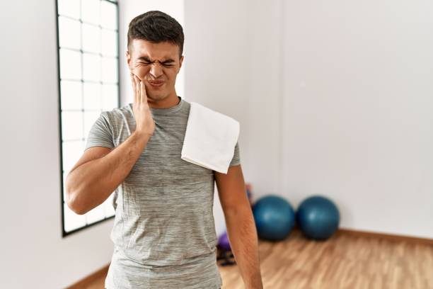 junger hispanischer mann, der sportbekleidung und handtuch im fitnessstudio trägt und den mund mit der hand mit schmerzhaftem ausdruck wegen zahnschmerzen oder zahnerkrankungen an den zähnen berührt. zahnarzt - dentist pain human teeth toothache stock-fotos und bilder