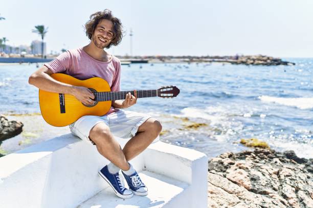 istock Young hispanic man playing classical guitar sitting on the bench at the beach. 1333977248