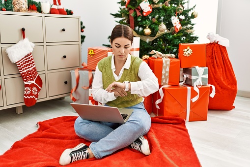 Young beautiful hispanic woman using laptop sitting by christmas tree checking the time on wrist watch, relaxed and confident