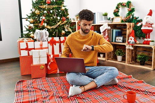 Arab young man using laptop sitting by christmas tree looking at the watch time worried, afraid of getting late