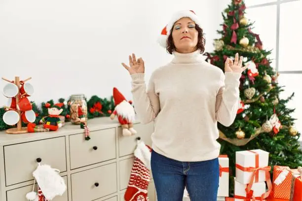Photo of Middle age hispanic woman standing by christmas tree relax and smiling with eyes closed doing meditation gesture with fingers. yoga concept.