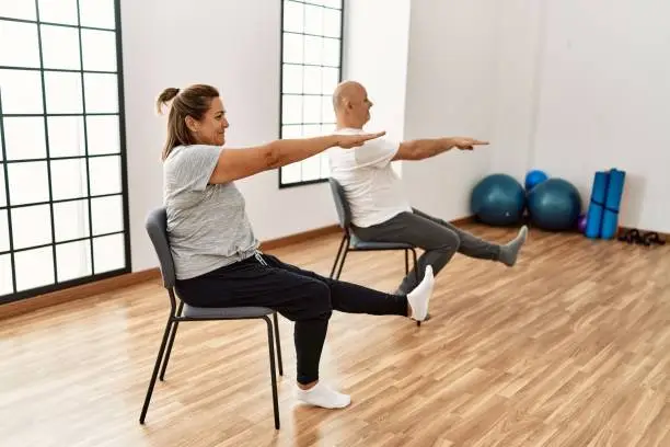 Photo of Middle age hispanic couple stretching using chair at sport center.