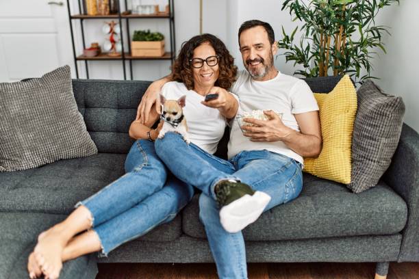 中年のヒスパニック系のカップルが映画を見たり、自宅のソファに座ってポップコーンを食べたりします。 - senior women electronics people remote control ストックフォトと画像