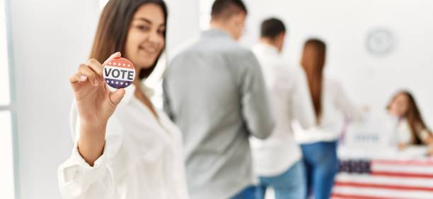 gruppe junger amerikanischer wähler, die ihre stimme abgeben. frau, die glücklich lächelt und das abzeichen im wahlzentrum in der hand hält. - voting stock-fotos und bilder