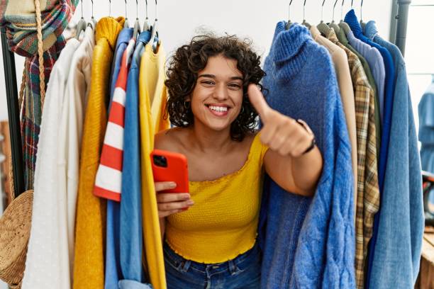 istock Young hispanic woman searching clothes on clothing rack using smartphone approving doing positive gesture with hand, thumbs up smiling and happy for success. winner gesture. 1333987537