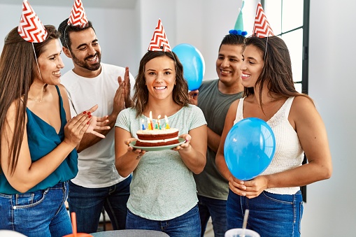 Young hispanic woman smiling happy and holding birthday cake standing with a group of friends at home.