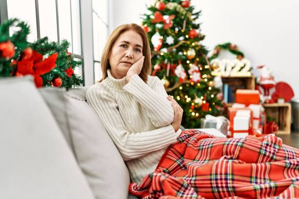 donna di mezza età seduta sul divano accanto all'albero di natale pensando di sembrare stanca e annoiata da problemi di depressione con le braccia incrociate. - senior women depression sadness women foto e immagini stock