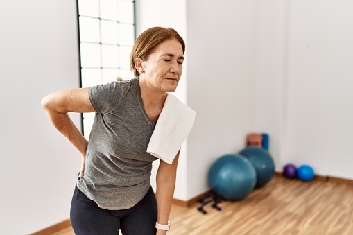 Middle age woman wearing sporty look training at the gym room suffering of backache, touching back with hand, muscular pain