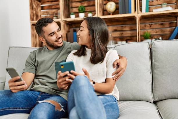 istock Young latin couple using smartphone sitting on the sofa at home. 1333989622