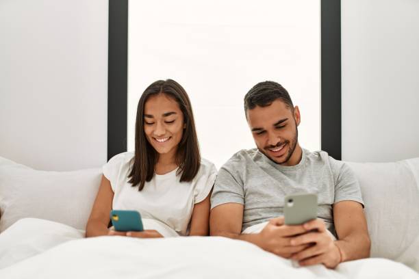 istock Young latin couple using smartphone lying on bed at home. 1333989687