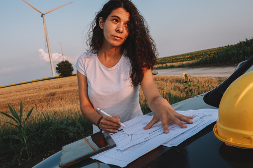 Female scientist working in the field, inspecting wind turbine efficiency. She is checking blueprints and real time inspection via digital tablet.