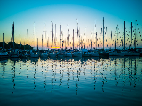 Sailboats are moored in the harbor with the sun setting in the background. Boats and masts reflect in the water. Postcard from Istria, Croatia. Summer.