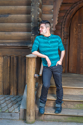 Dressing in a green striped long sleeves Henley shirt, black pants and leather boots, a young handsome guy is standing outside a wooden house and taking a break.\