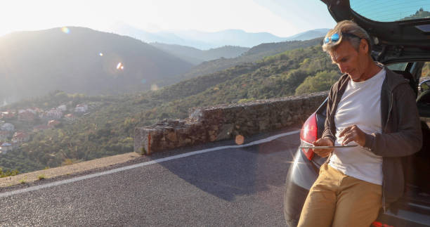 istock Man relaxes against car on rural road, and uses tablet 1334011213