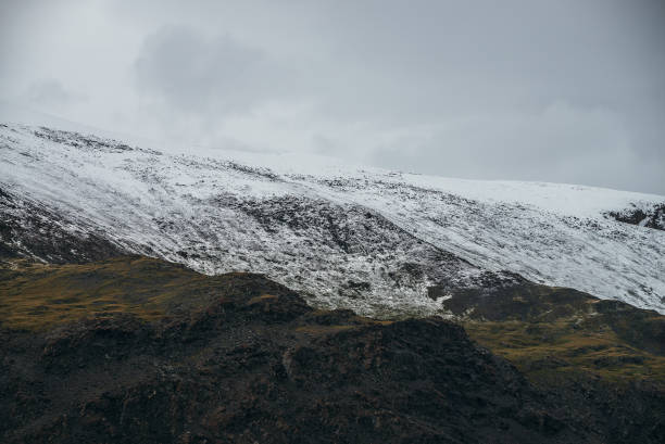 Minimalist highlands landscape with snow-capped mountains under cloudy sky. Snowbound high mountain range in overcast weather. Atmospheric minimalism with green mountains, white snow and gray sky.
