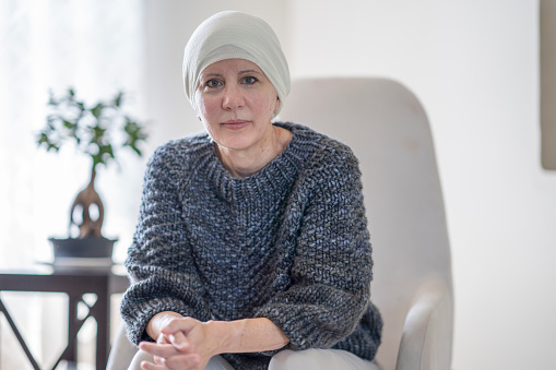 A mature Caucasian woman with cancer sits on her chair in her living room. She is wearing a white scarf on her head to cover her hair loss. She is looking at the camera with a somber expression on her face.