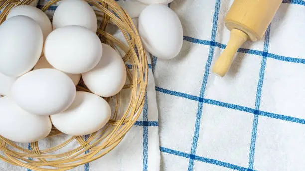 Close-up of a small bamboo basket with white eggs on blue checkered tablecloth, a rolling pin, top view